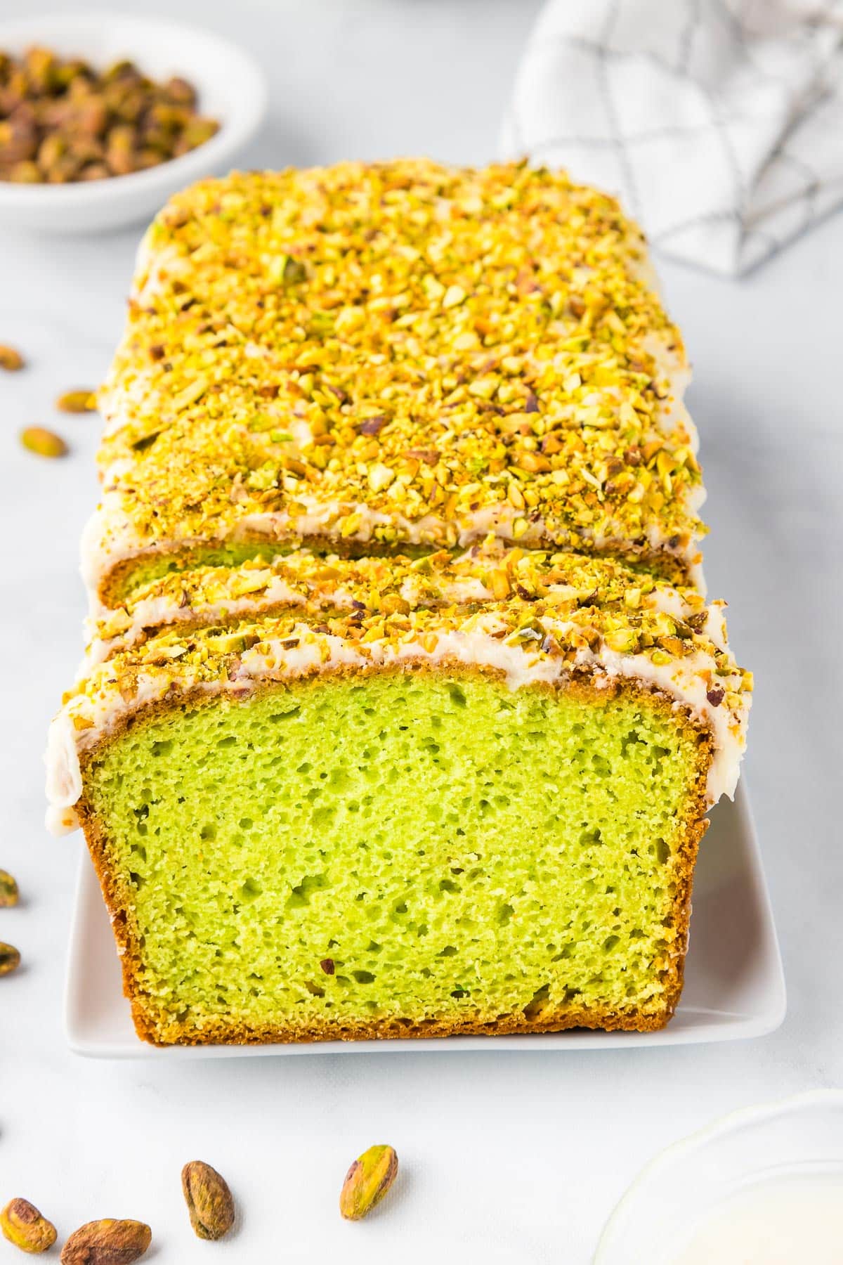 A sliced loaf of green pistachio bread with glaze and crushed pistachios on top on a counter with more pistachios in around the bread and in a bowl.