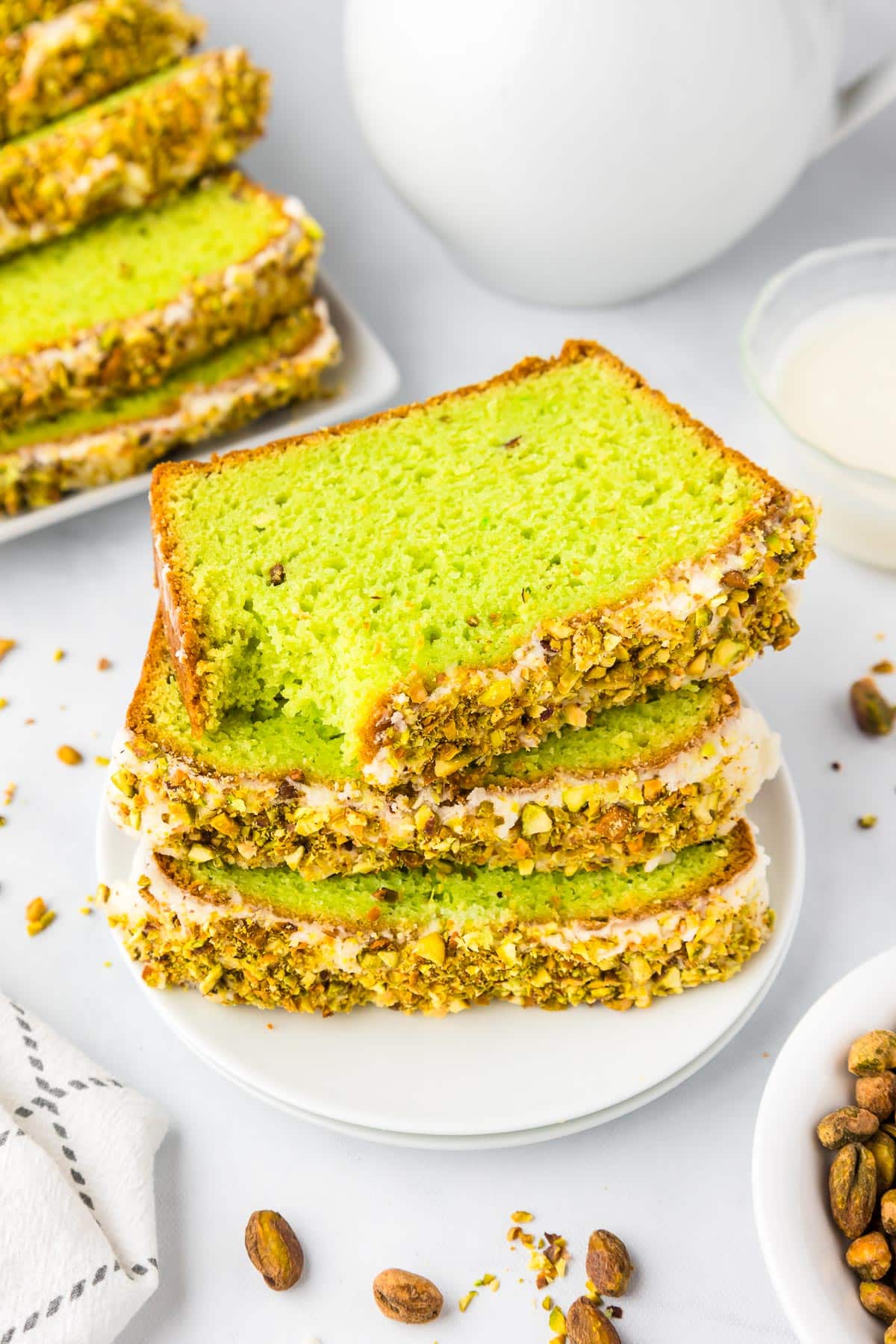 Three slices of green pistachio cake with sweet glaze and chopped pistachio stacked on a white plate with the top piece missing a bite, and more pistachio bread on a serving dish on the counter nearby.