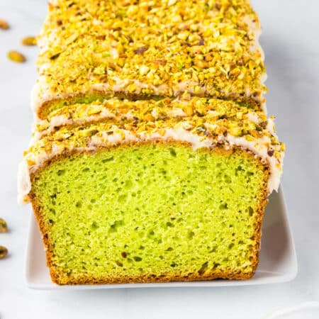 Square view of a sliced loaf of pistachio bread with green interior and white glaze, topped with chopped pistachios, on a white plate.