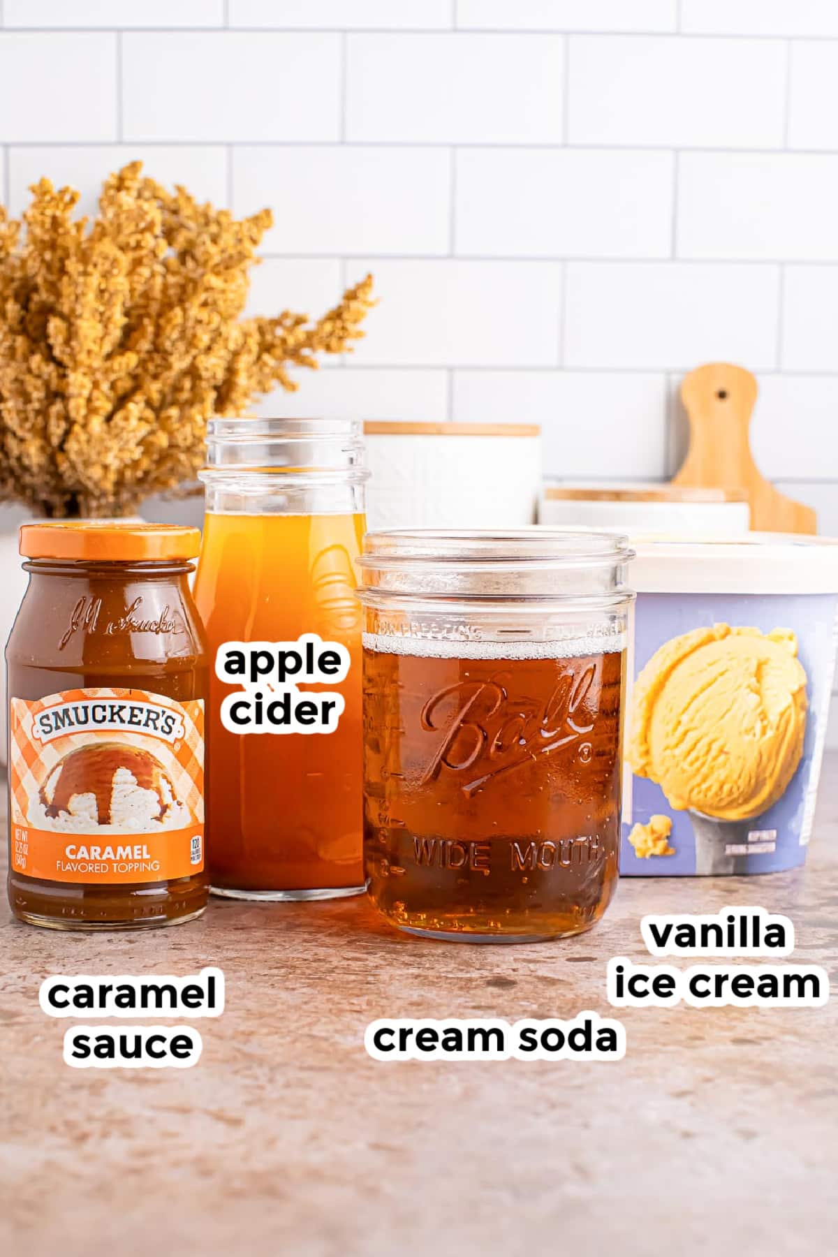 Ingredients for an apple cider float including apple cider, cream soda, ice cream and caramel sauce on a counter with text labels.