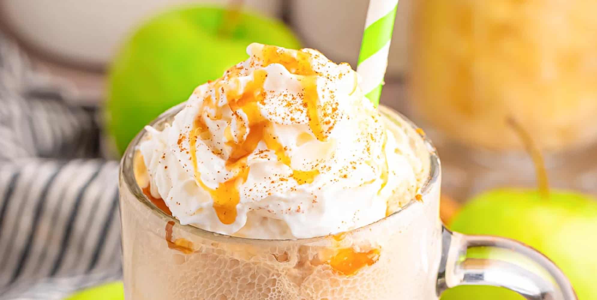 A wide view of a glass mug filled with an apple cider float, topped with whipped cream, caramel drizzle, and a green-striped straw. Green apples and another drink are in the background.