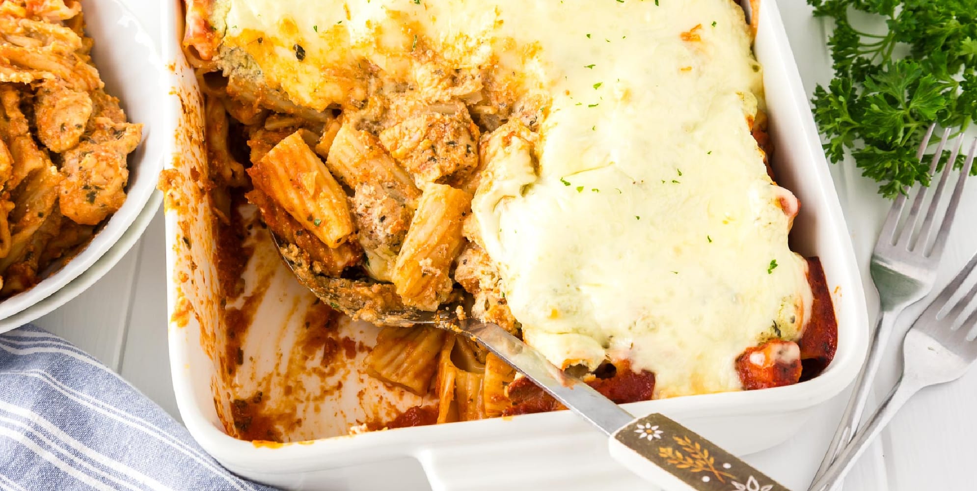 Baked rigatoni with chicken in a casserole dish from above with a portion missing and in a bowl near the baking dish and a serving spoon in the pan.