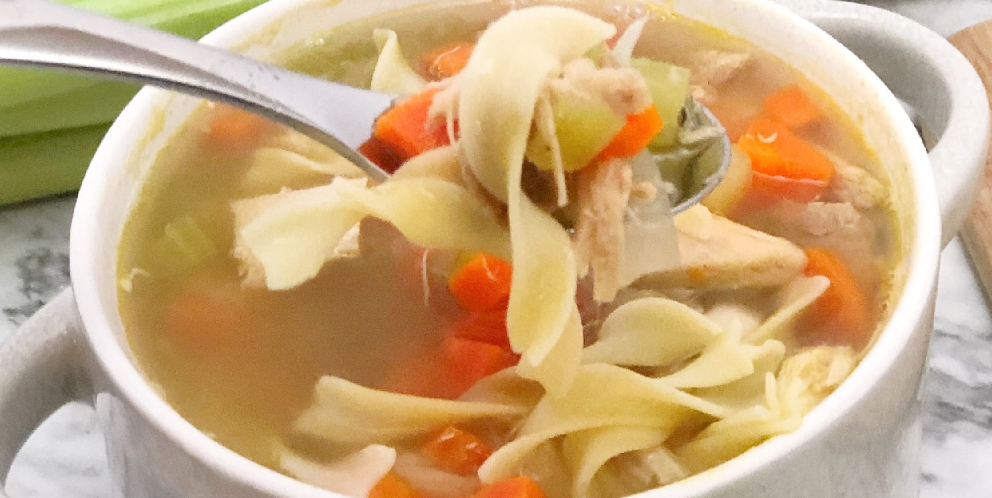 Close-up of a bowl of chicken noodle soup with a spoon lifting a portion. The soup contains noodles, carrots, celery, and chicken pieces.