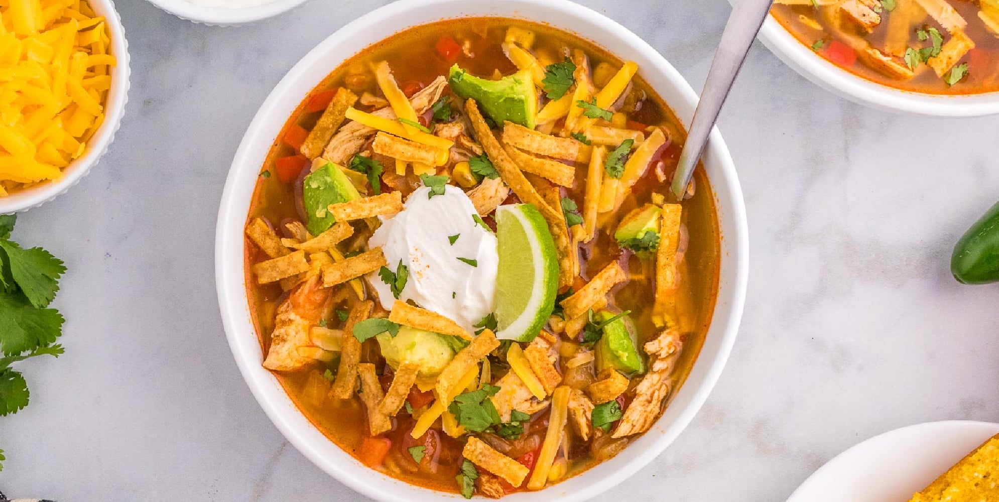 A bowl of chicken tortilla soup topped with shredded cheese, tortilla strips, avocado, lime wedge, and a dollop of sour cream, with a spoon inside with other bowls of ingredients on the counter nearby.