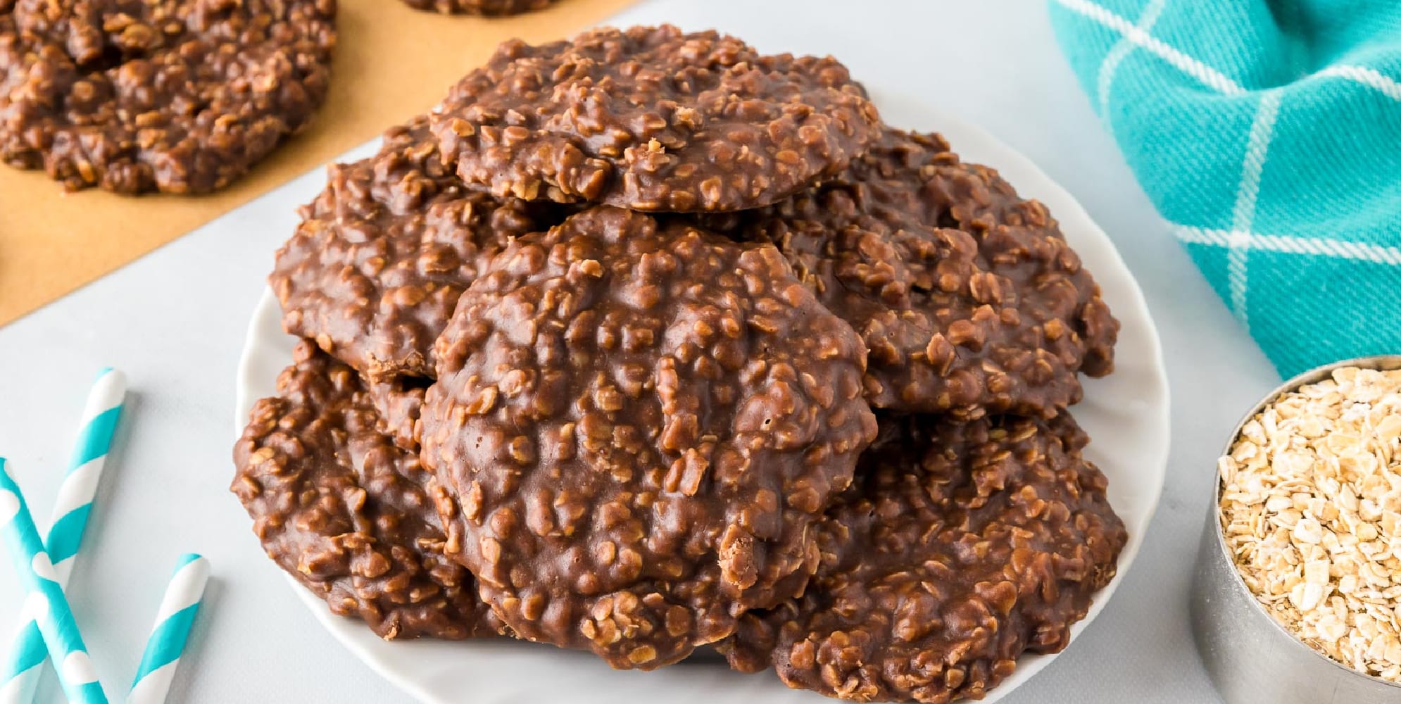 A wide view of a plate piled with several no-bake chocolate peanut butter oatmeal cookies with more cookies nearby on parchment paper.