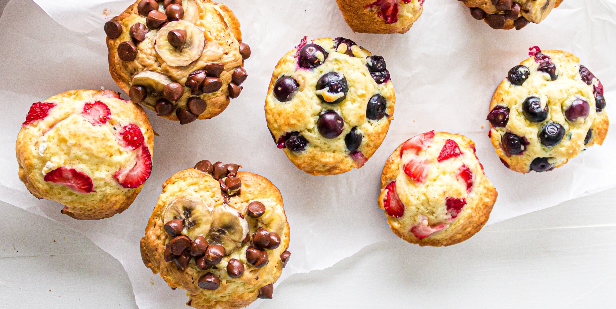 A variety of muffins with different toppings such as strawberries, chocolate chips and banana slices with chocolate chips on parchment paper from above,