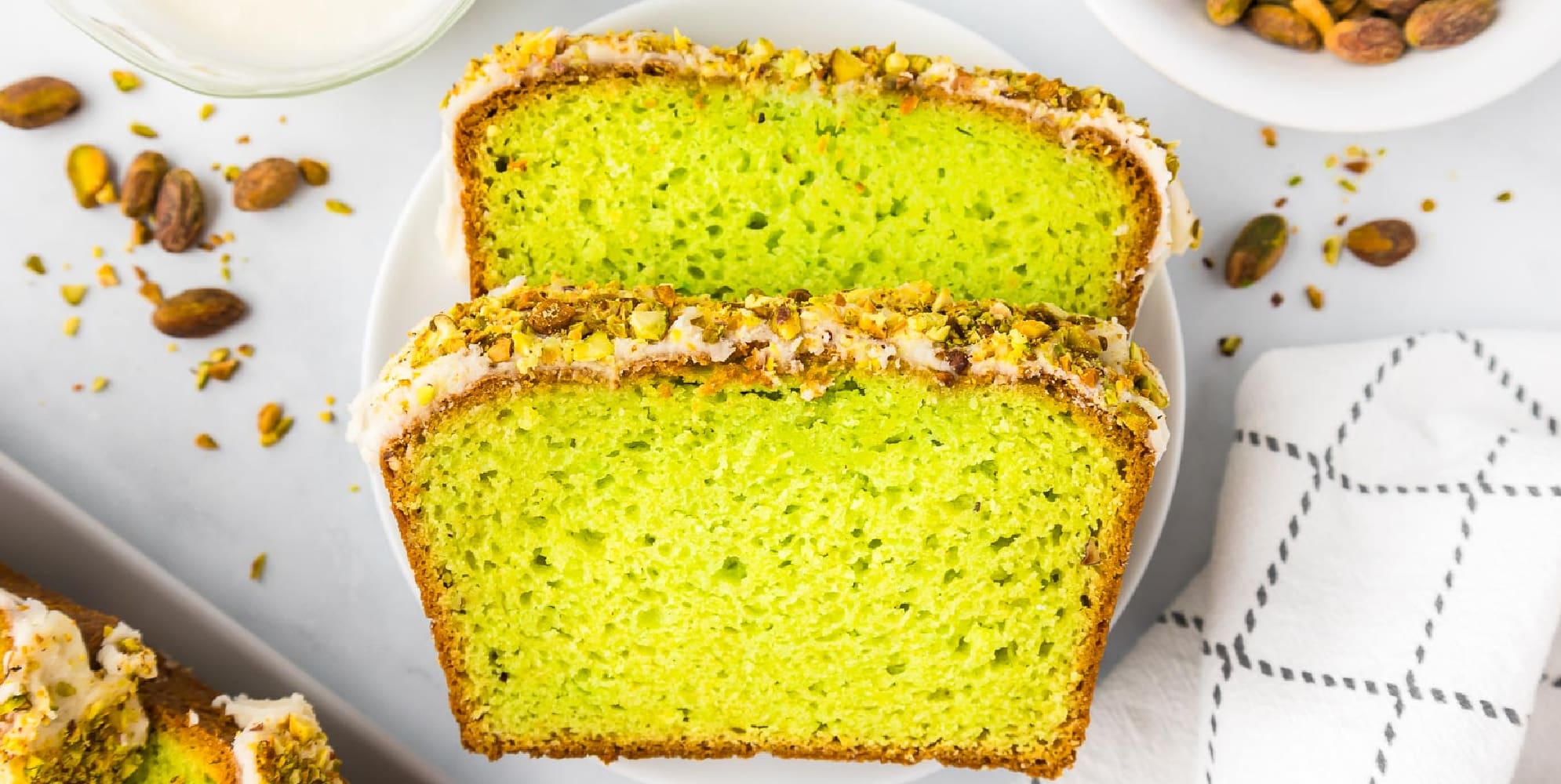 Two slices of bright green pistachio cake topped with white glaze and chopped pistachios on a white plate from above with chopped pistachios and more pistachio bread in a pan nearby.