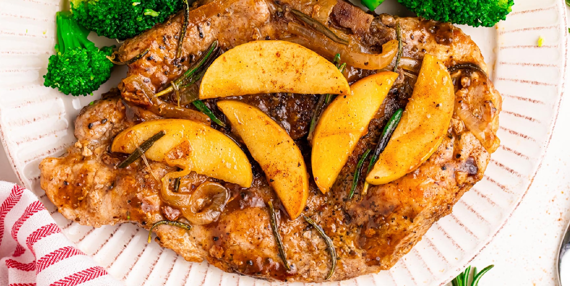 Wide view of a cooked apple butter pork chip on a plate with cooked apples and onions on top next to steamed broccoli on the plate.