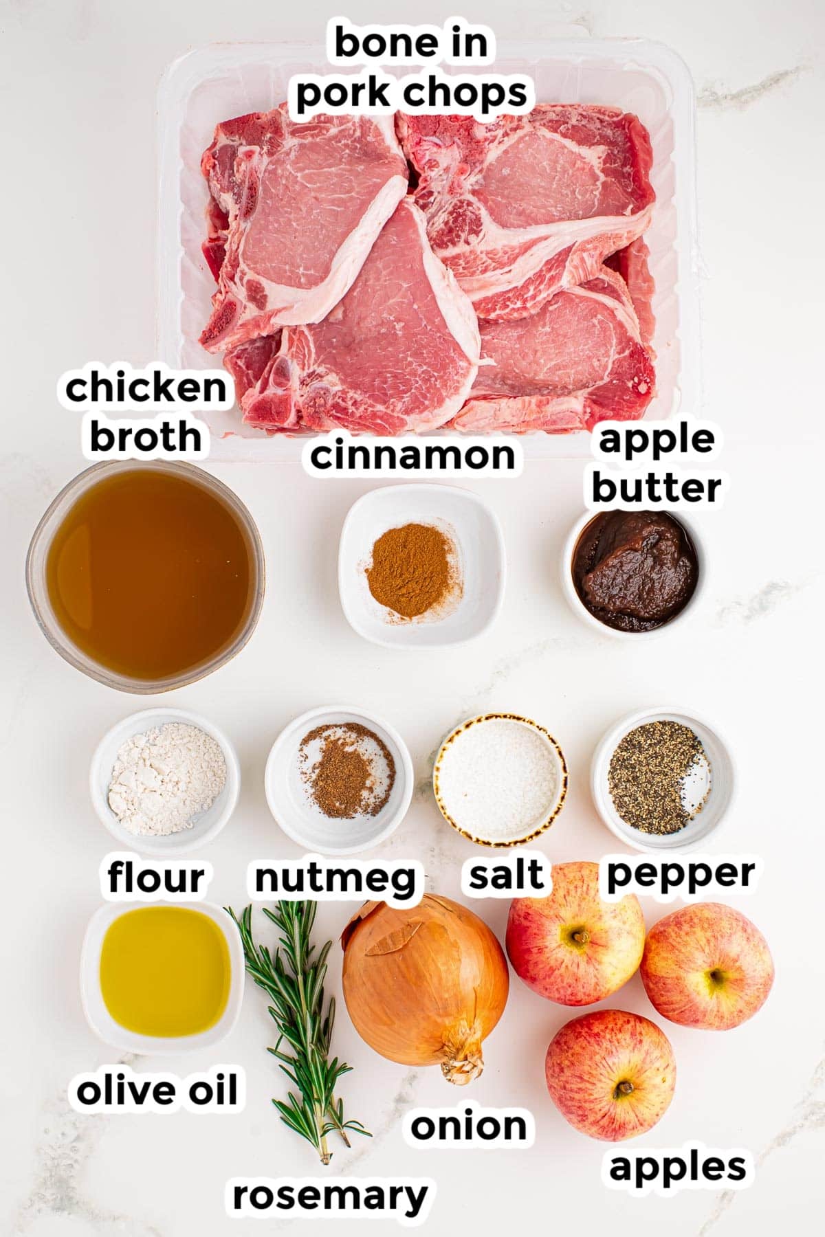 Top-down view of bone-in pork chops, chicken broth, cinnamon, apple butter, flour, nutmeg, salt, pepper, olive oil, rosemary, onion, and apples arranged on a white surface with labels.