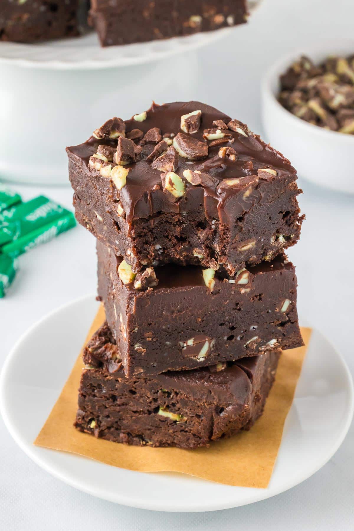 A close-up of three chocolate Andes mint brownies stacked on plate with a thick ganache frosting and topped with andes mint pieces. The top brownie is missing a bite.