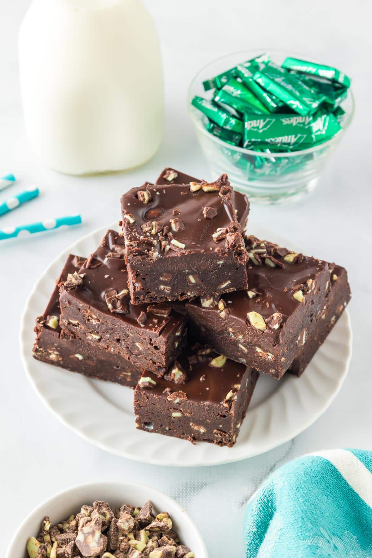 A plate stacked high with fudgy Andes Mint brownies topped with mint pieces, next to a glass bowl with green-wrapped mints and a glass bottle of milk and a bowl of more foil wrapped Andes mints.