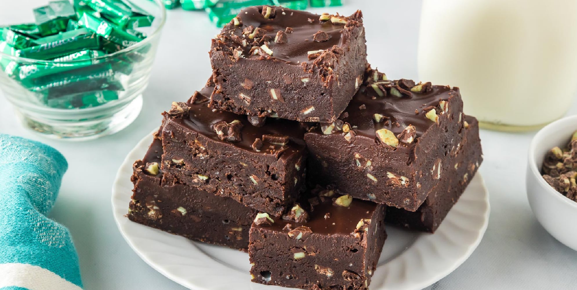 A white plate holds a stack of chocolate brownies topped with chopped nuts. Mint candies and a glass of milk are in the background. A blue and white towel is to the left of the plate.