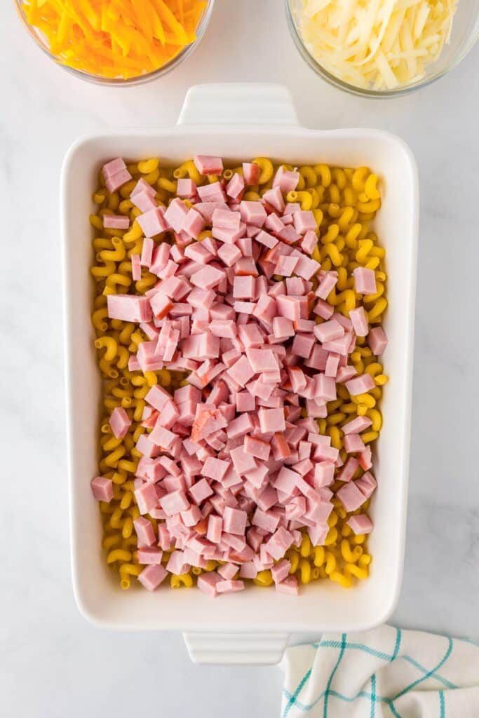 An overhead view of uncooked cavatappi pasta in a rectangular baking dish for mac and cheese being mixed with diced ham with cheese in bowl nearby.