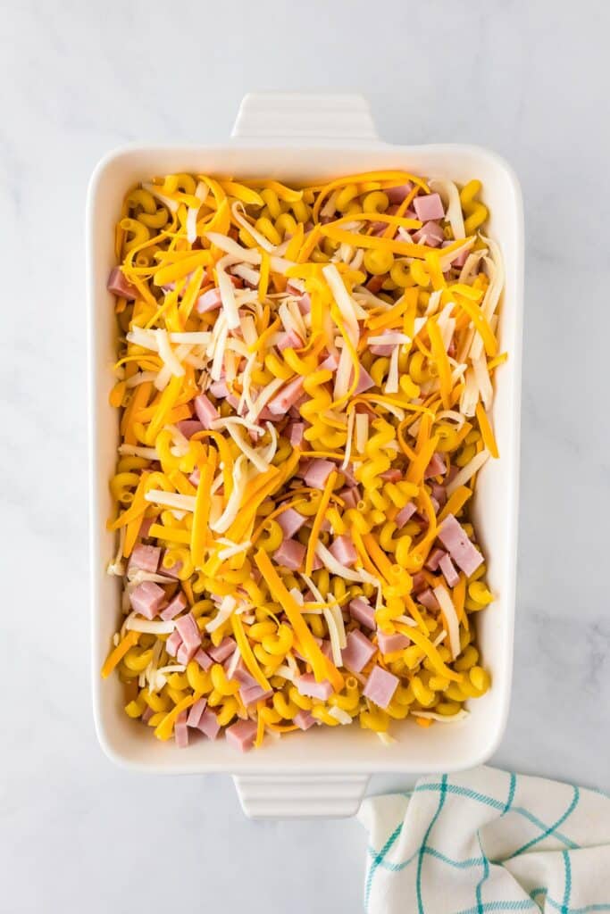 An overhead view of uncooked cavatappi pasta in a rectangular baking dish for mac and cheese being mixed with diced ham and a lot of shredded cheese.