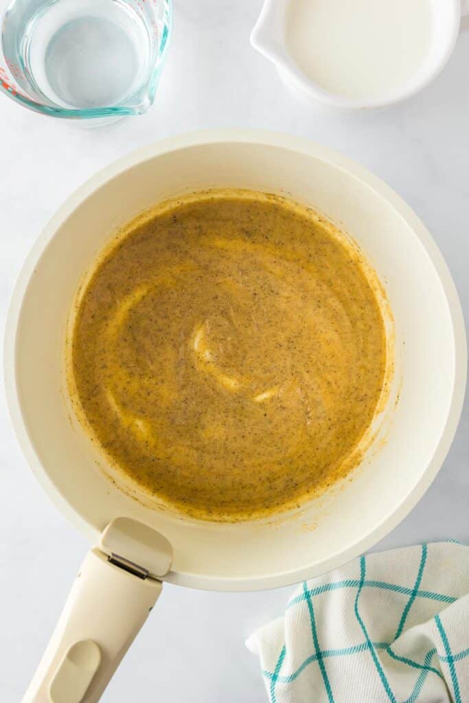 A saucepan containing melted butter, spices and flour cooking to make a roux for cavatappi mac and cheese with water and milk in bowls nearby.