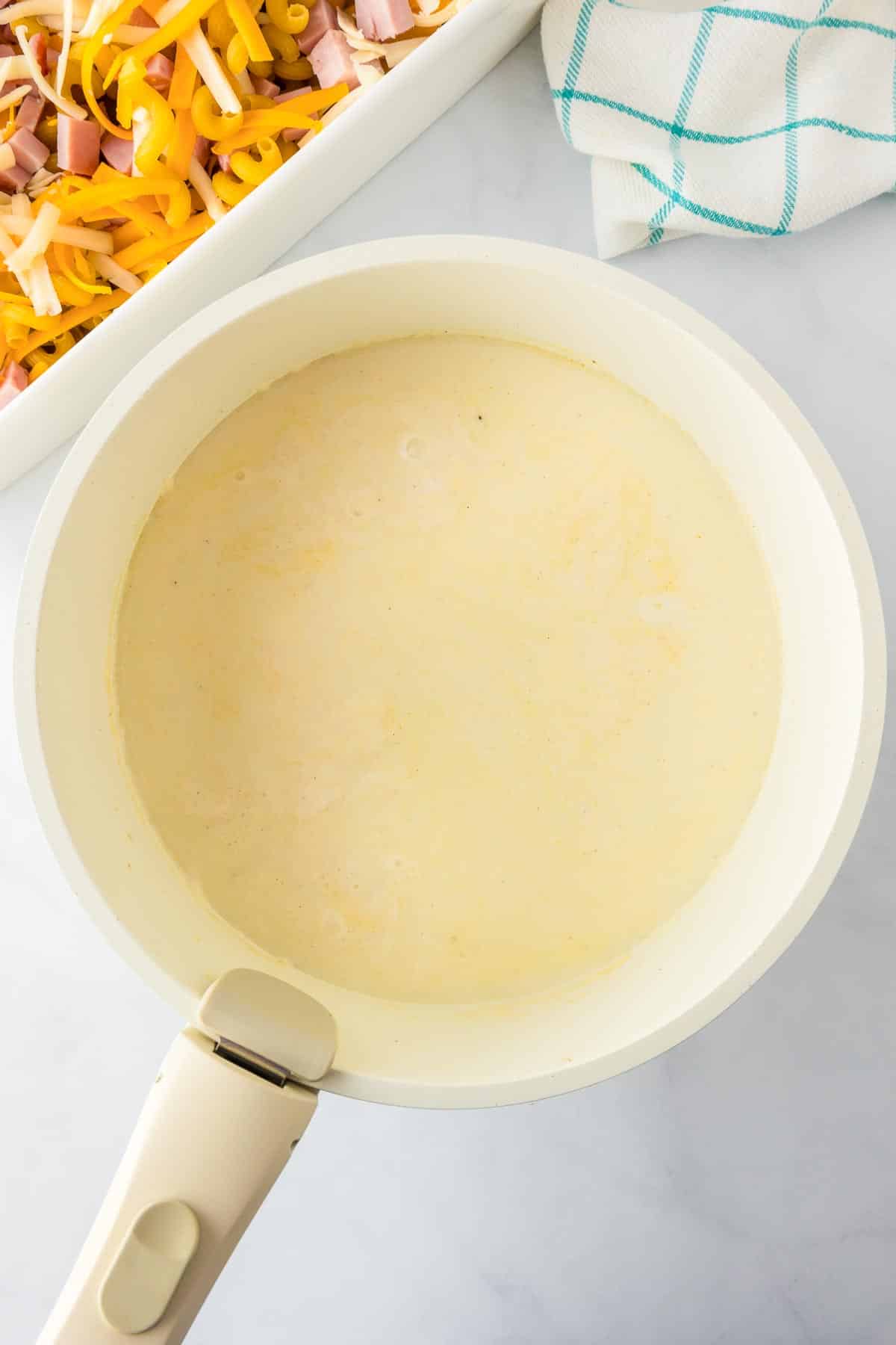 A pot with a creamy sauce next to a casserole dish filled with uncooked cavatappi pasta, ham and cheese.