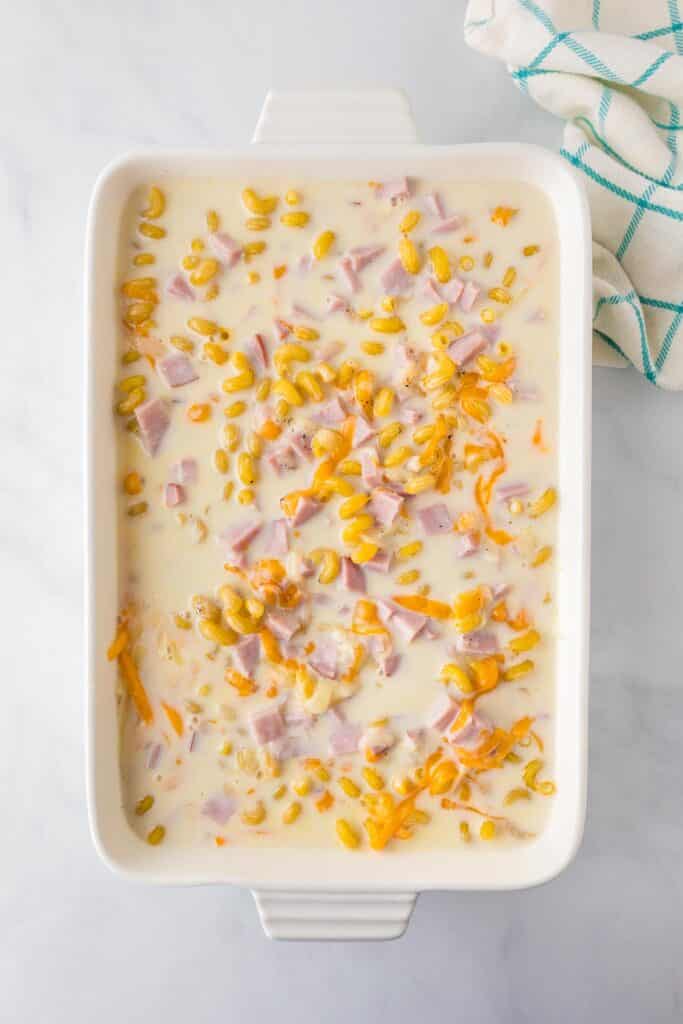 A white rectangular baking dish filled with raw cavatappi noodles, ham, and shredded cheese in a milky sauce for baked cavatappi mac and cheese.