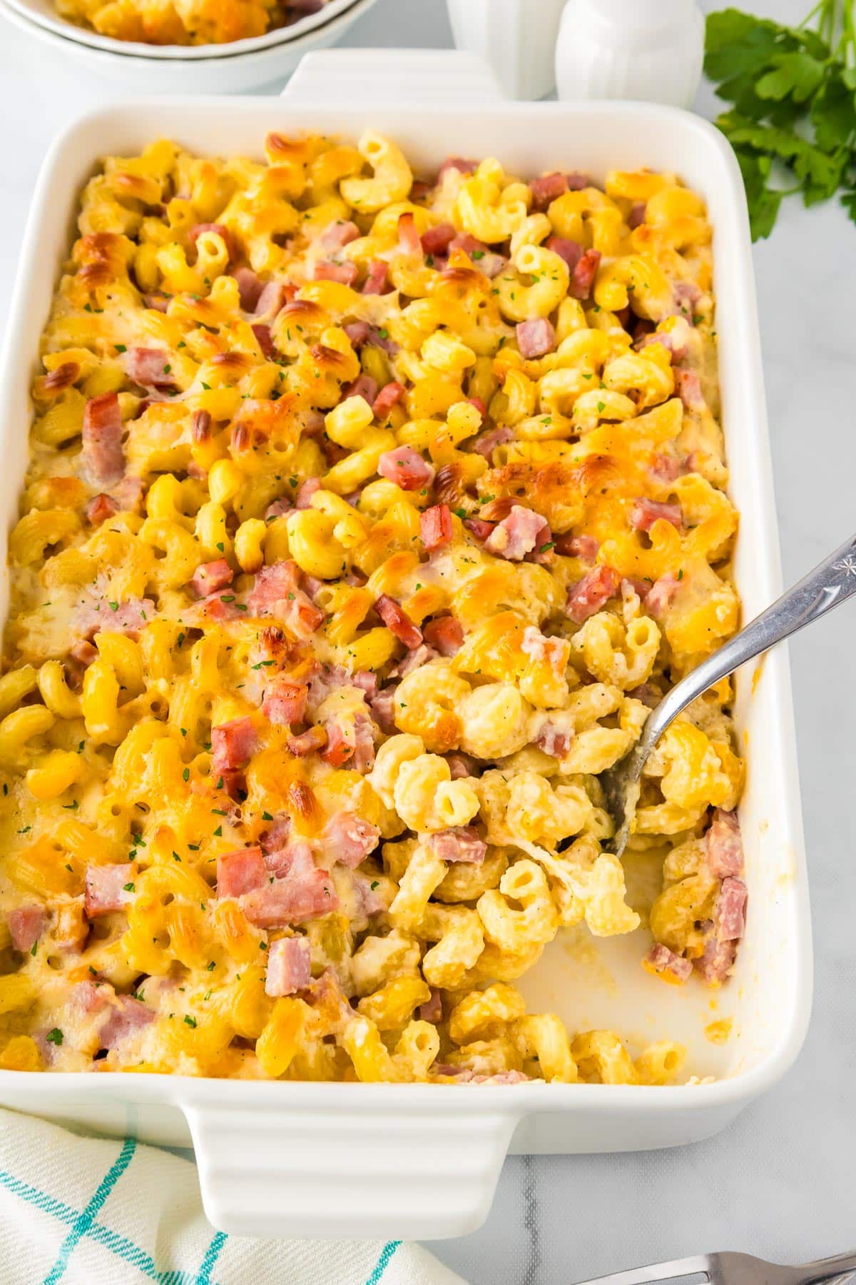 A baked cavatappi macaroni and cheese casserole with diced ham being served in a white rectangular dish with a spoon.