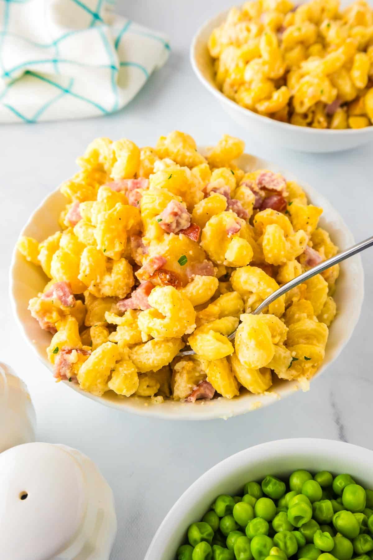 Bowl of corkscrew mac and cheese with ham with more mac and cheese in a second bowl and a bowl of peas nearby.