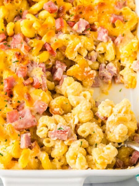 A square view of a serving pan filled with baked cavatappi macaroni and cheese topped with diced ham and melted cheese while a spoon scoops the pasta.