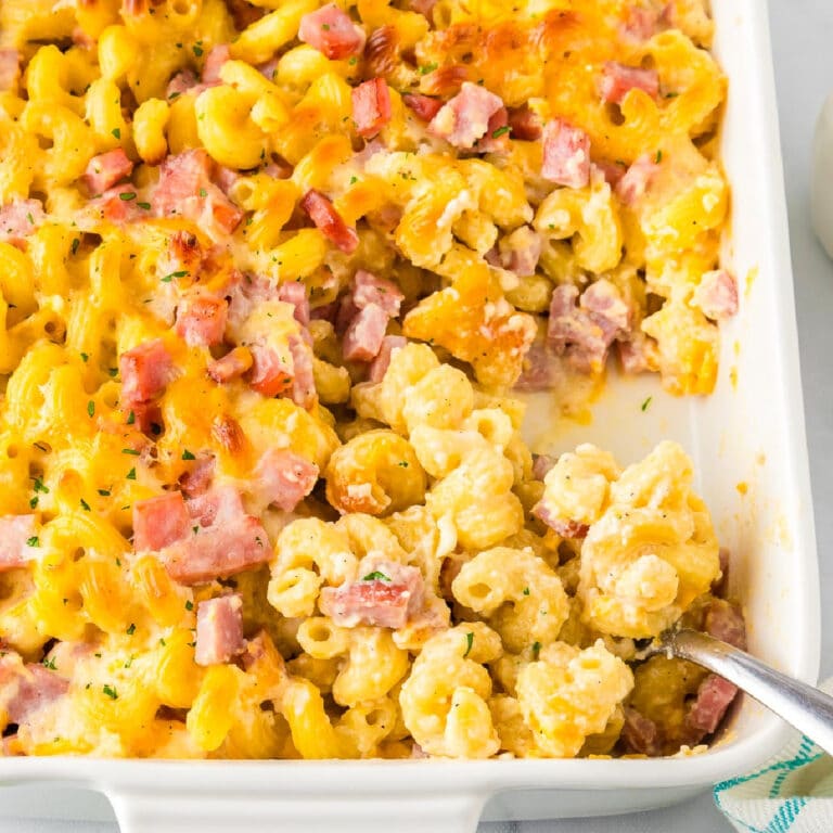 A square view of a serving pan filled with baked cavatappi macaroni and cheese topped with diced ham and melted cheese while a spoon scoops the pasta.