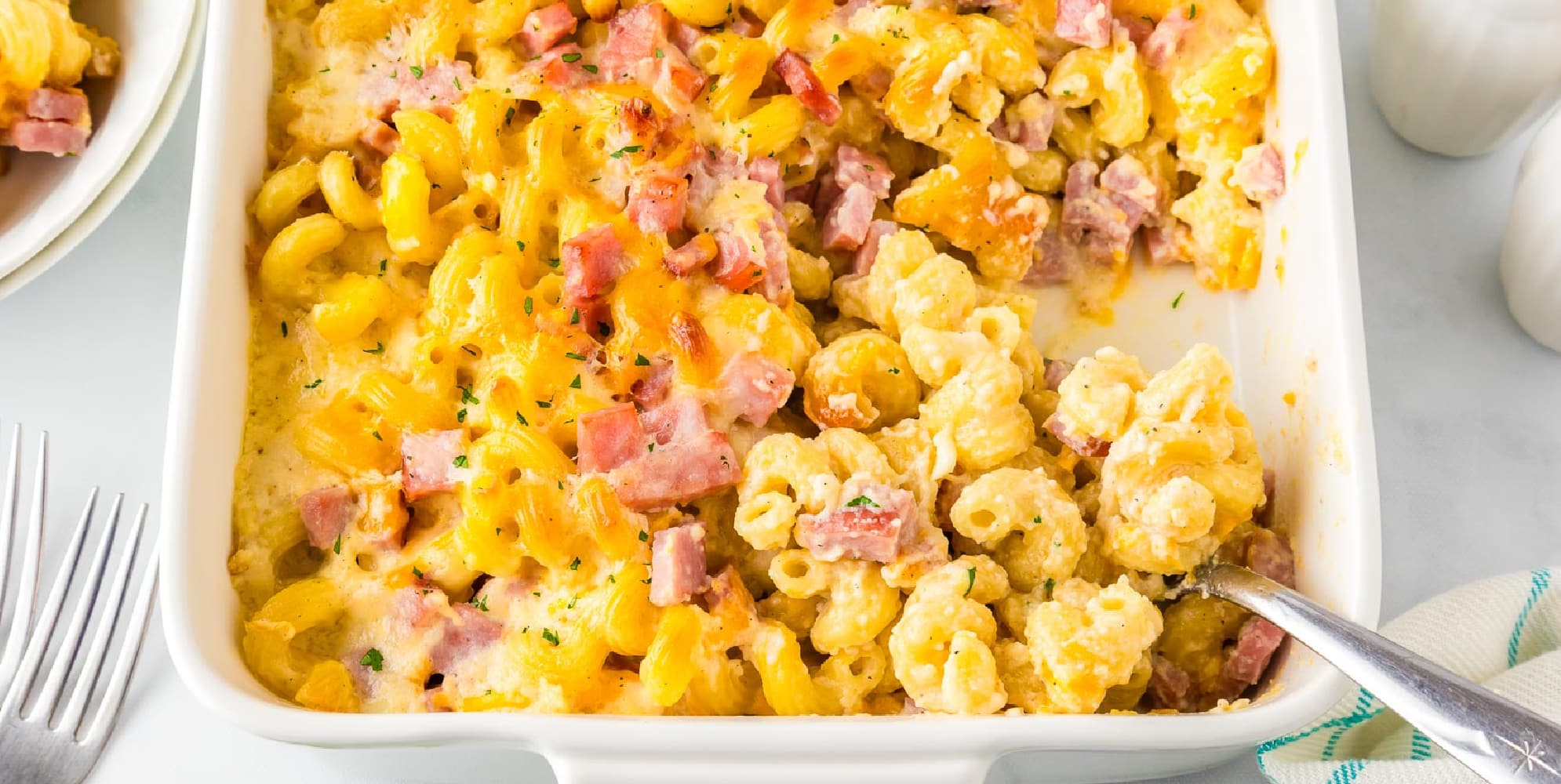 A wide view of a casserole dish with baked cavatappi macaroni and cheese mixed with ham, partially scooped out to reveal the creamy interior texture.