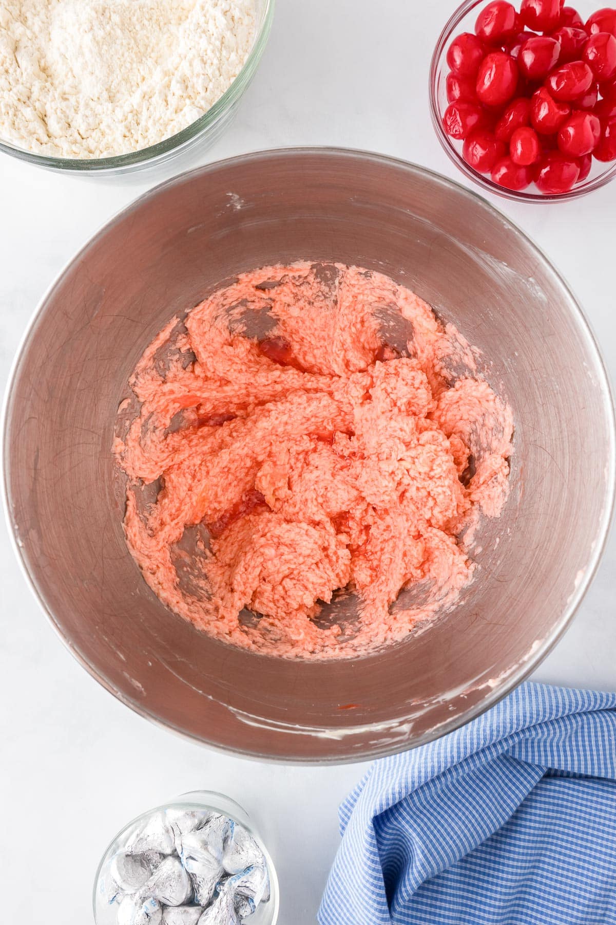 A mixing bowl with pink cookie dough next to bowls of flour, cherries, chocolate kiss candies for cherry kiss cookies.