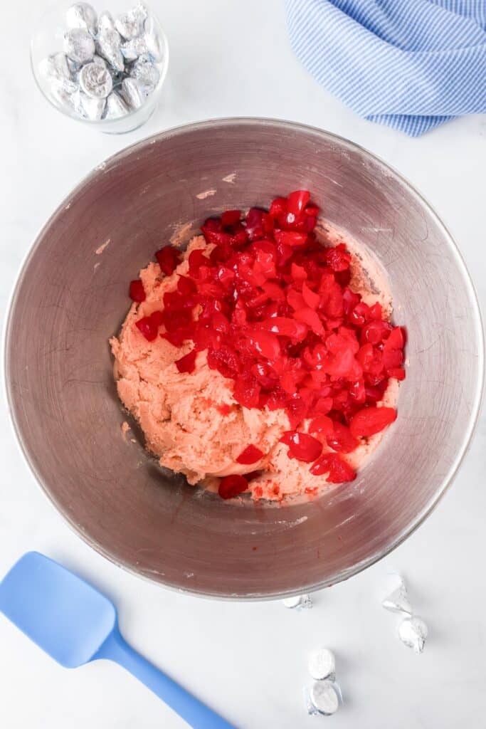 Mixing bowl with pink cookie dough and chopped red cherries being folded in with a silicone spatula and chocolate kisses nearby on the counter.