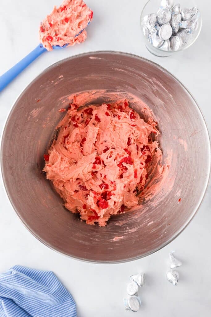 A bowl of pink cookie dough for cherry kiss cookies with red maraschino cherry pieces folded in, a silicone spatula and a bowl of chocolate kisses nearby on the counter.