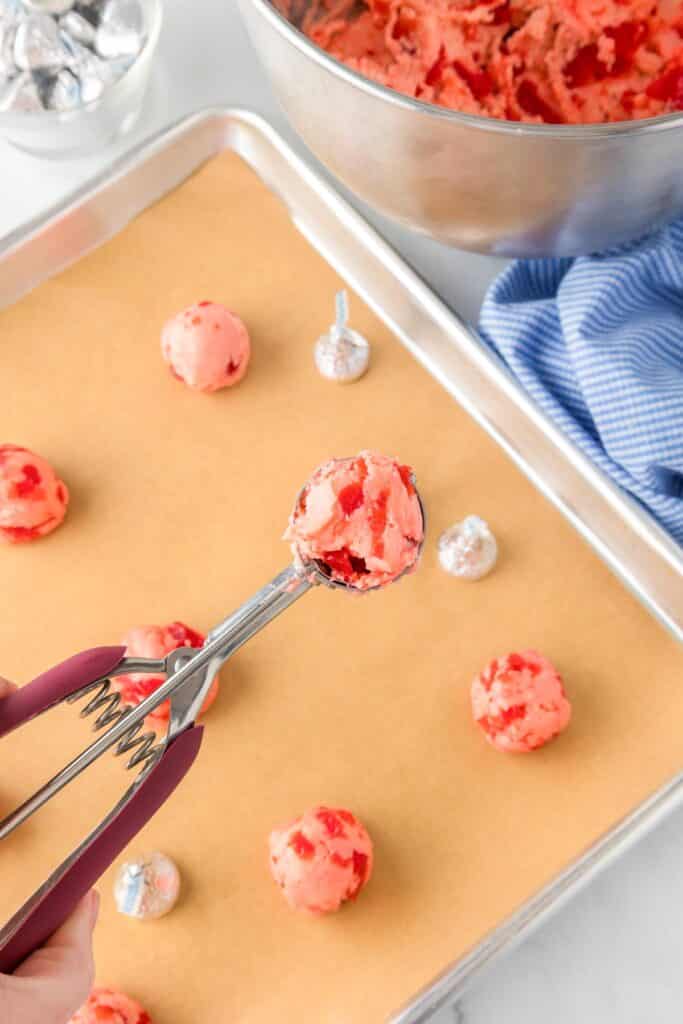 A cookie scoop holds cherry kiss cookie dough with more cookie dough balls on a pan with a bowl of dough nearby.