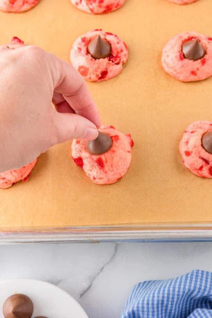 A hand places a chocolate kiss piece on a pink cherry kiss cookie on a cookie sheet next to other cookies.