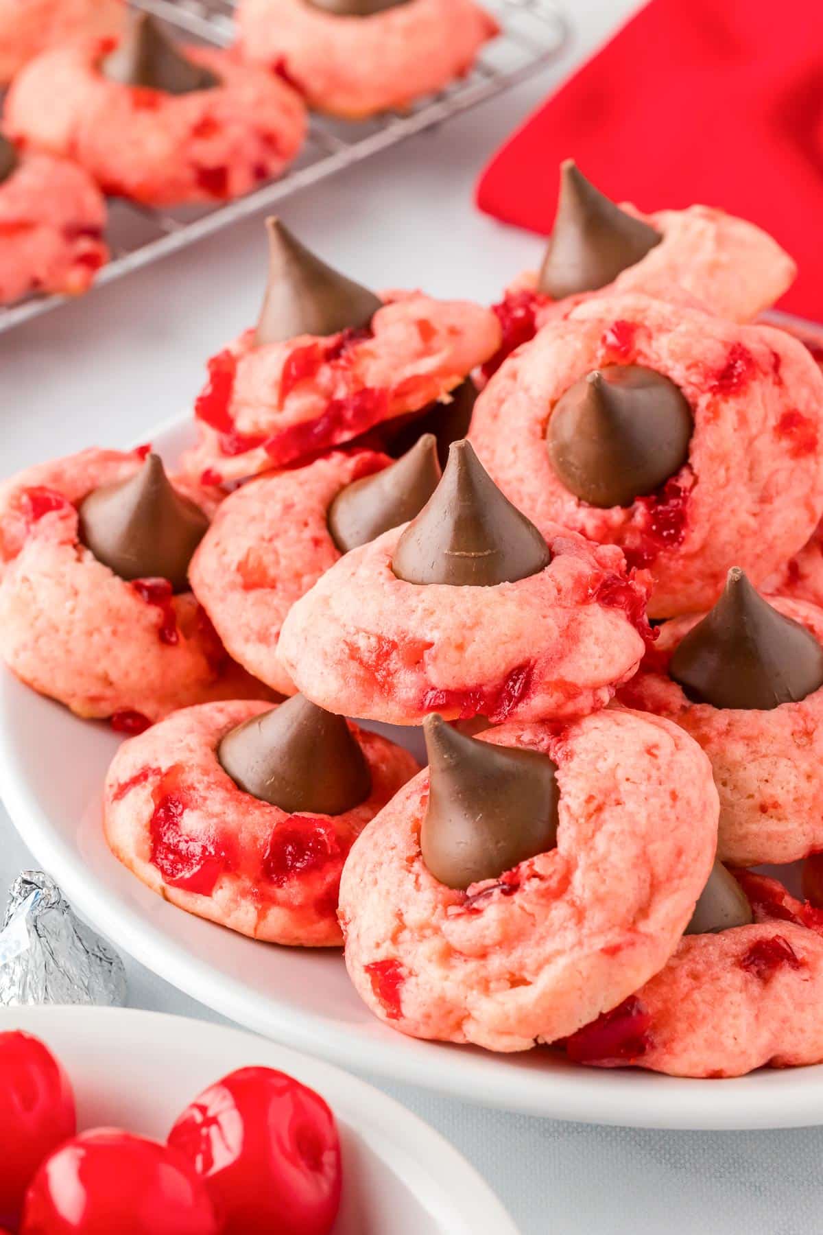 A plate of pink and red cherry kiss cookies topped with chocolate kisses on a platter piled high with more cookies on a wire rack in the background.