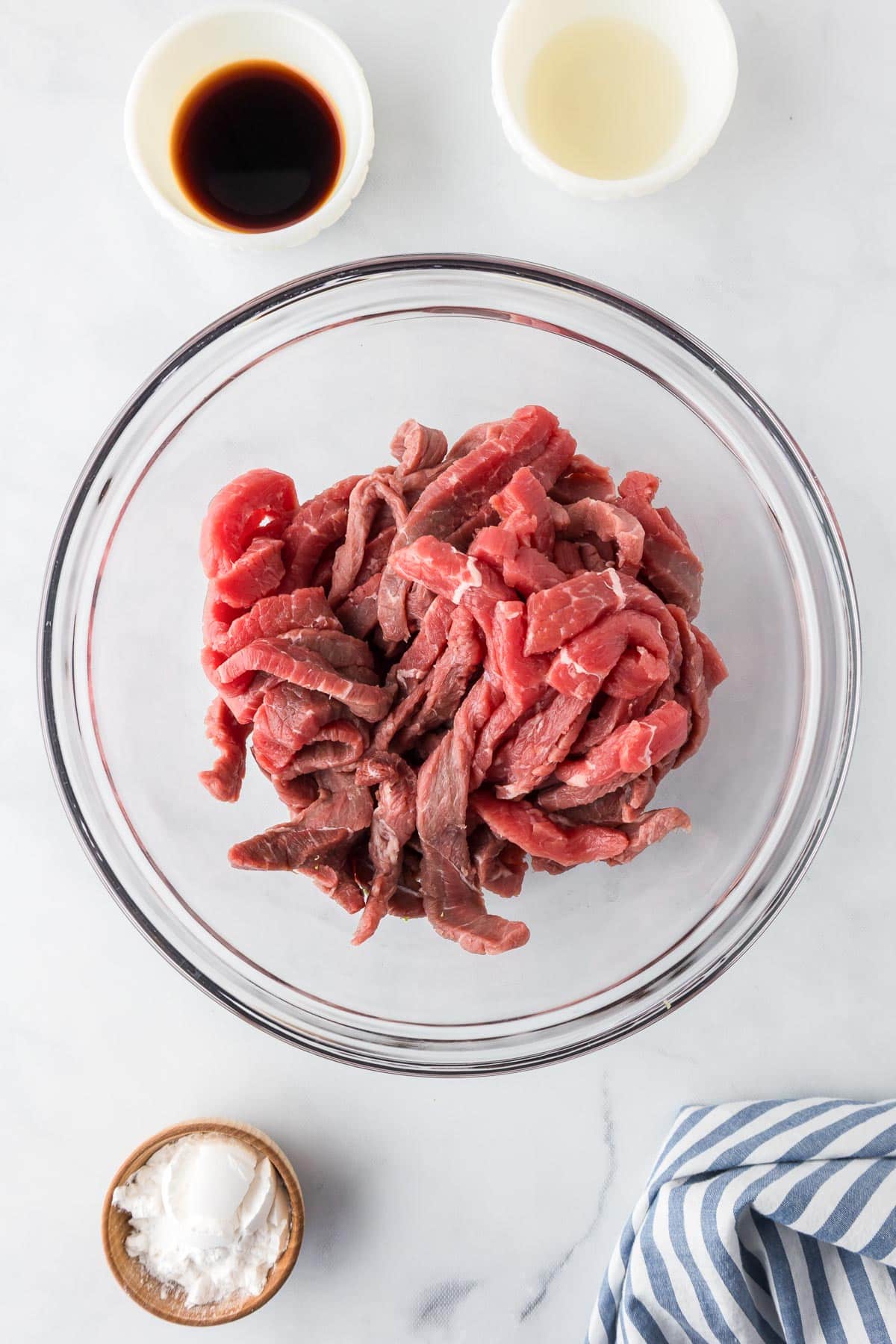 A bowl of raw thin sliced steak with small bowls of cornstarch, soy sauce and rice wine vinegar nearby.