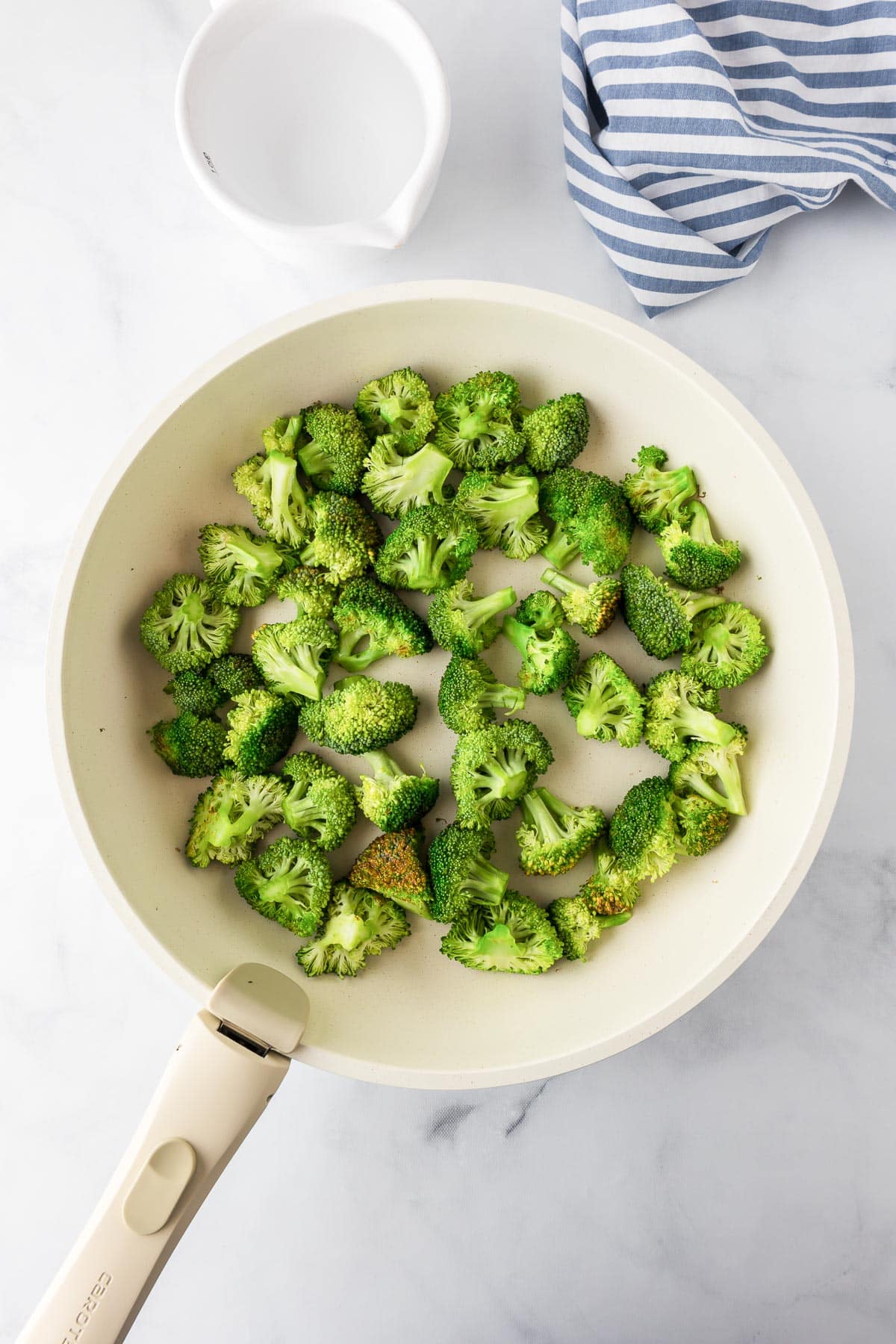 A pan with bright green cooked broccoli florets with water in a cup nearby.