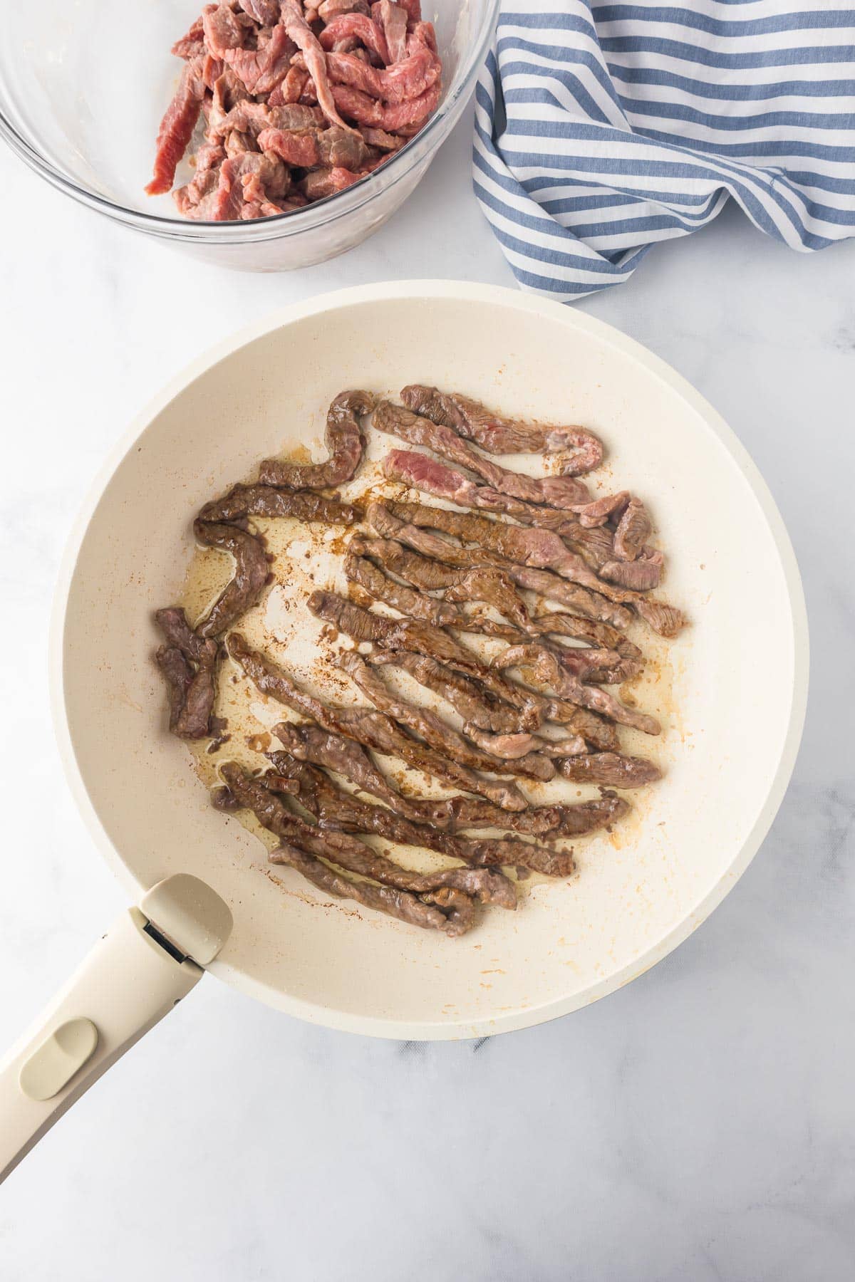 Slices of beef cooking in a pan until golden brown with more raw thin sliced broccoli in a bowl nearby.