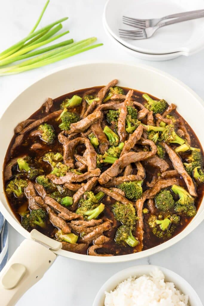 A finished skillet with beef and broccoli in a dark sauce with plates with forks, green onions and a bowl of steamed rice on the counter nearby.