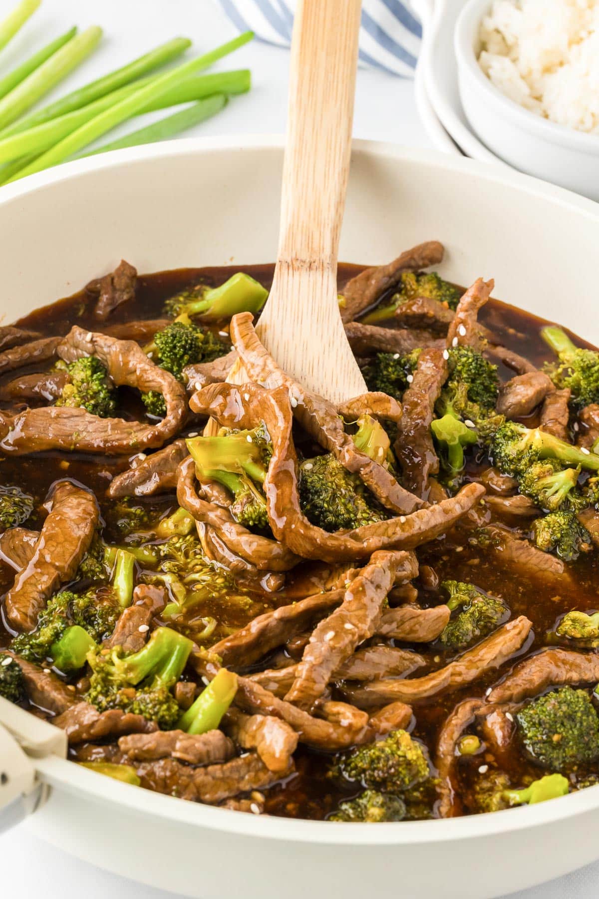 Beef with broccoli in a pan with a wooden spoon stirring it, and steamed rice in a bowl nearby.