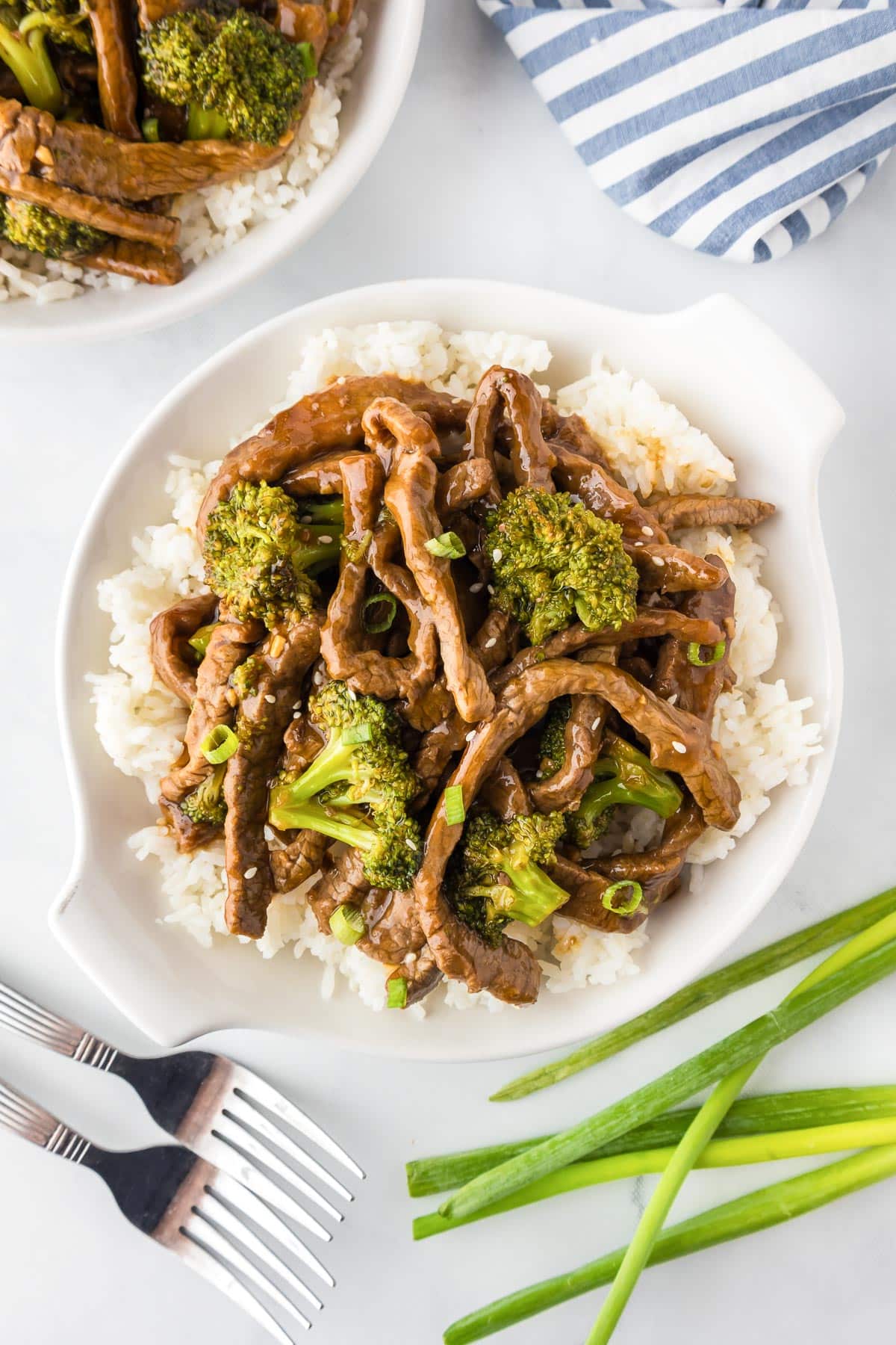 A plate of beef and broccoli stir-fry on white rice, garnished with green onions. Two forks and green onion with a second plate, a napkin and two forks nearby.