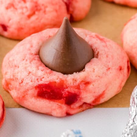 A pink cherry kiss cookie with a chocolate kiss in the center placed on a piece of parchment paper with more cookies in the background.