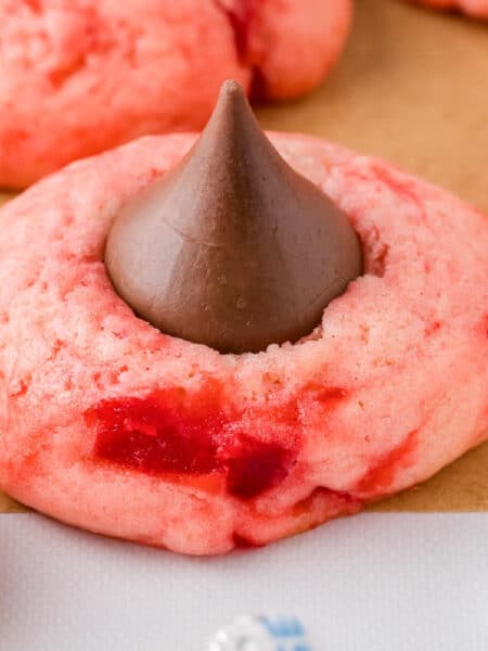 A pink cherry kiss cookie with a chocolate kiss in the center placed on a piece of parchment paper with more cookies in the background.