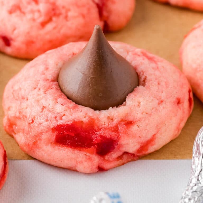 A pink cherry kiss cookie with a chocolate kiss in the center placed on a piece of parchment paper with more cookies in the background.