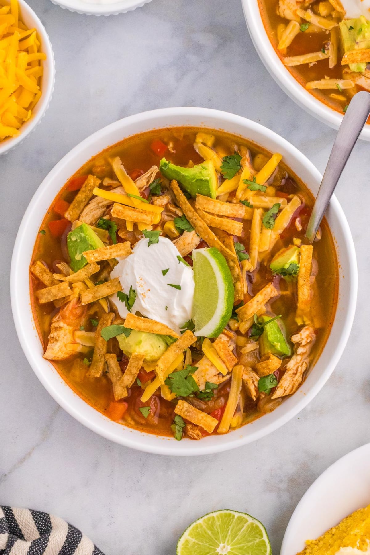 A bowl of chicken tortilla soup with avocado, tortilla strips, shredded cheese, sour cream, and a lime wedge on top on a counter from overhead.