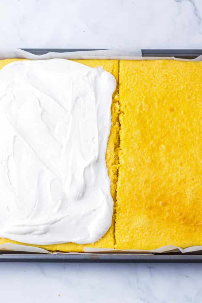 Yellow cake on parchment paper in a jelly roll pan cut in half with half of the cake being frosted with a marshmallow filling.