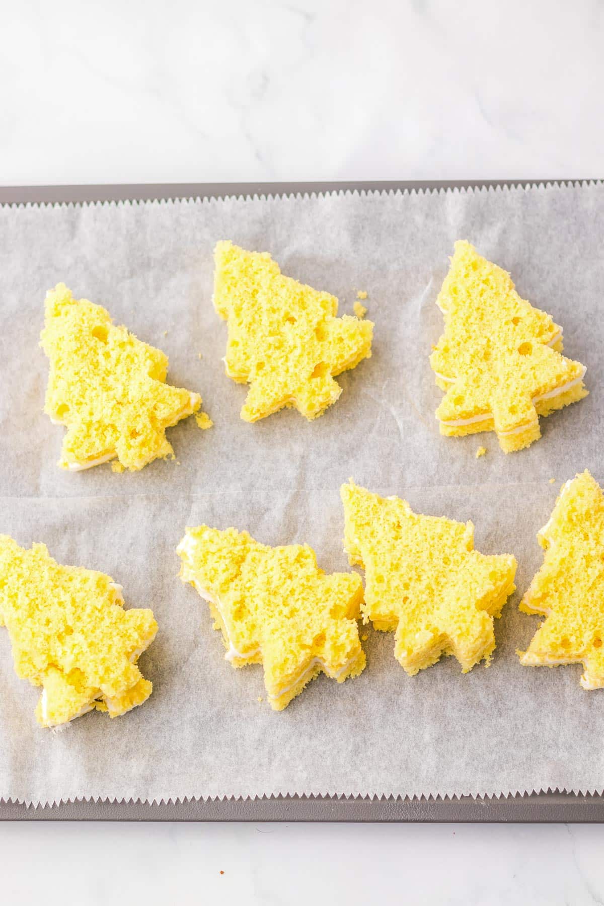 Seven tree-shaped yellow Christmas cakes filled with a marshmallow frosting on a parchment-lined baking sheet.