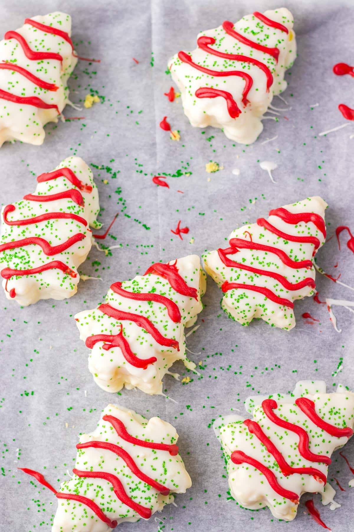 Tree-shaped cakes with white icing, red drizzle, and green sprinkles on parchment paper.