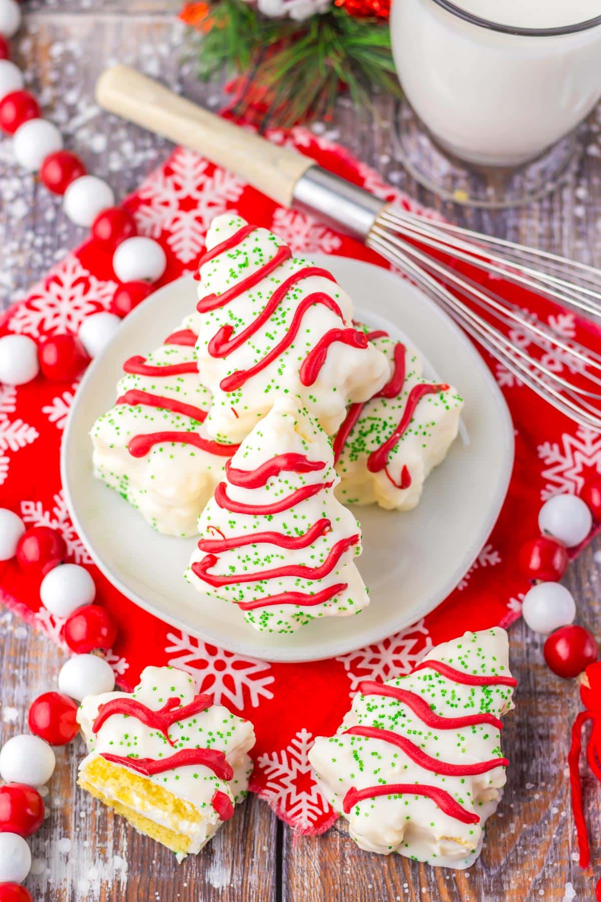 A stack of white copycat Little Debbie Christmas tree tree cakes with red icing and green sprinkles on a plate surrounded by a a glass of milk, a napkin and holiday decorations on the table.