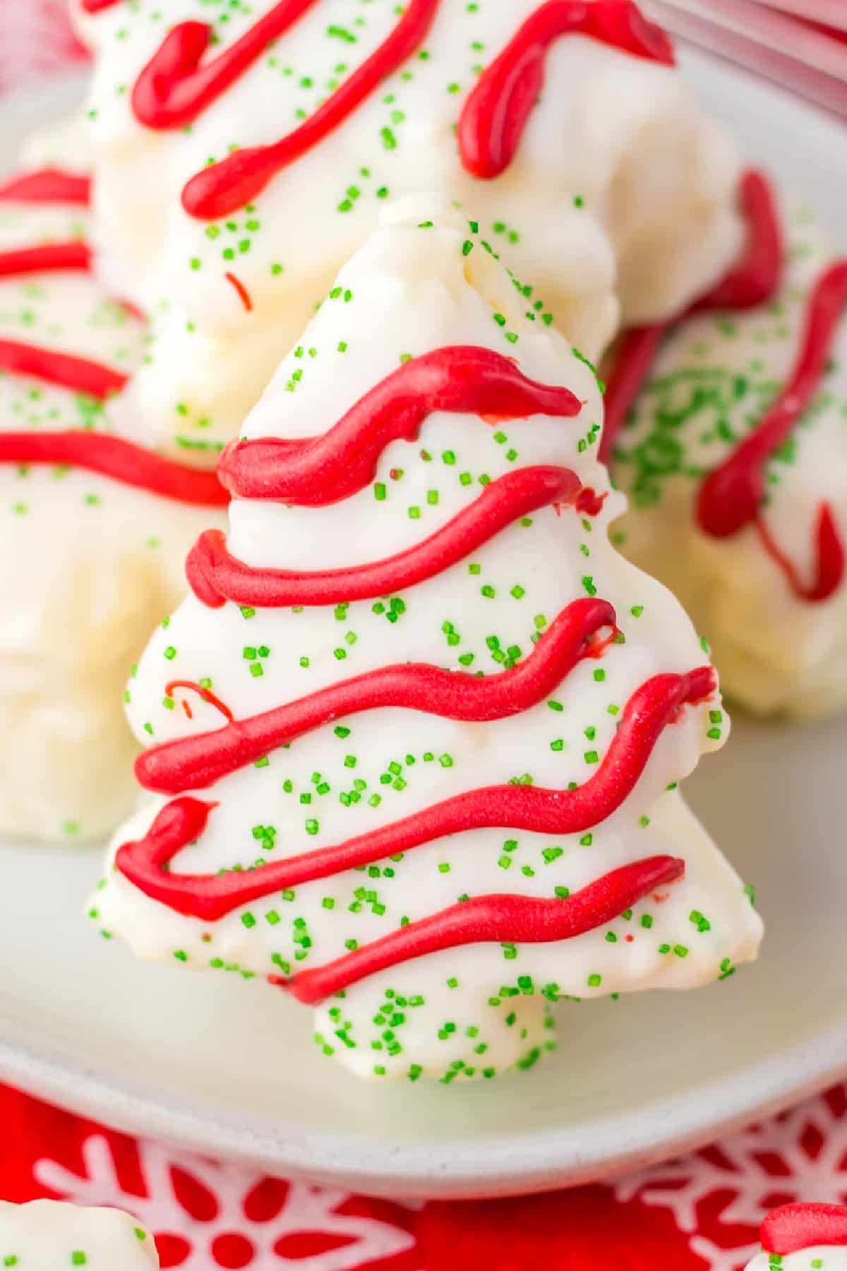 Copycat Christmas tree cake with white icing, red zigzag decorations and green sprinkles piled on a plate with more cakes.