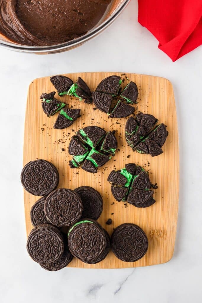 Mint Oreo cookies on a cutting board with several chopped roughly into four pieces to put in the nearby Grinch brownie batter in a bowl.