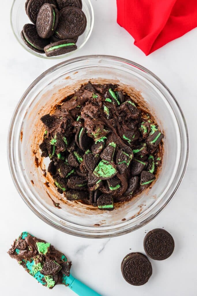 Mint Oreo cookie pieces being folded into a Grinch brownie batter in a bowl with a silicone spatula nearby.