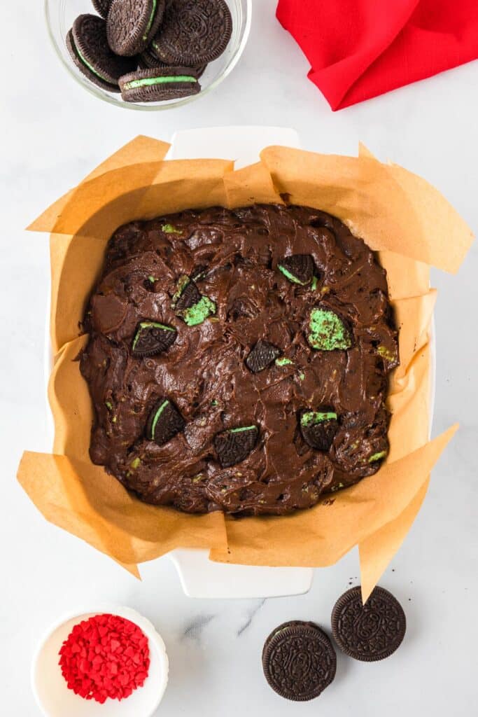 A pan of chocolate Grinch brownie dough with chunks of mint Oreo cookies with more cookies and red heart sprinkles nearby on the counter.